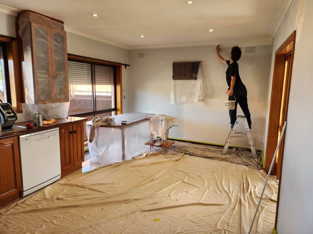 Painter cutting in along the cornice of a Melbourne kitchen, floor covered with drop sheets and cabinetry masked in plastic