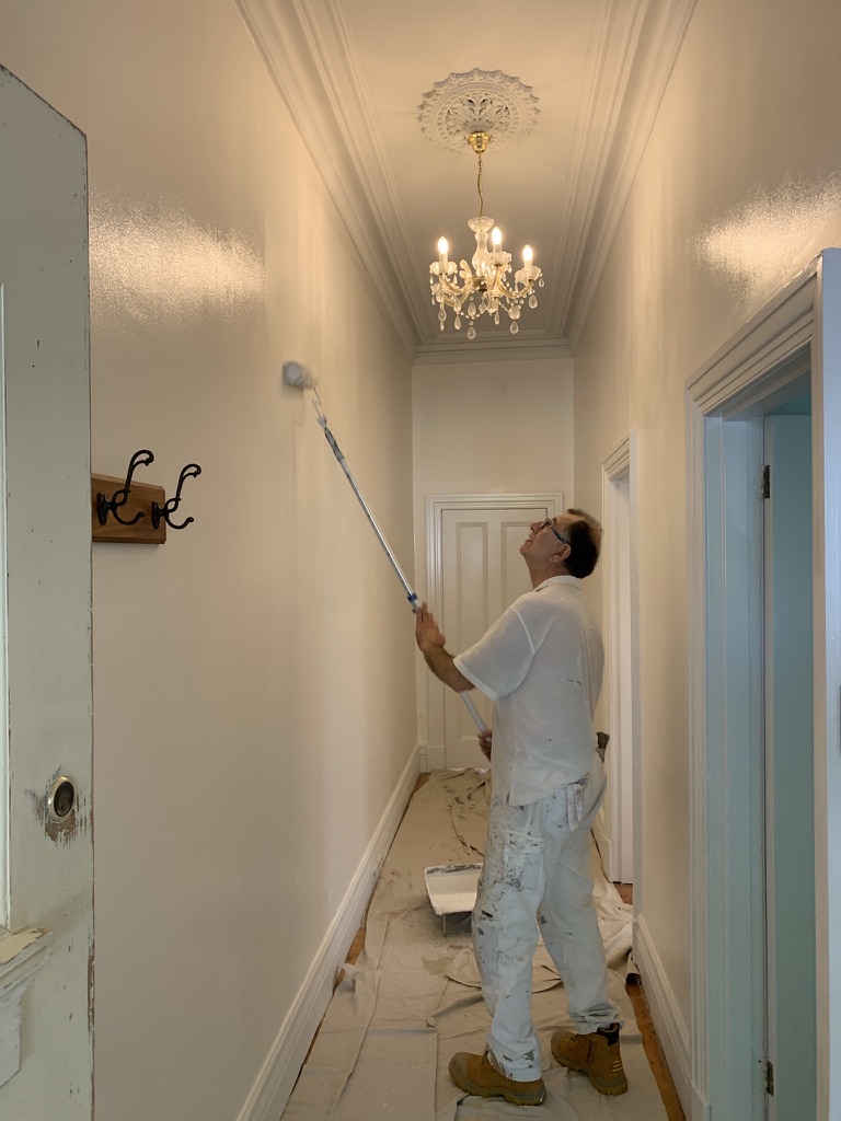 Interior painter rolling walls in a Brunswick terrace hallway — ornate ceiling rose and chandelier overhead