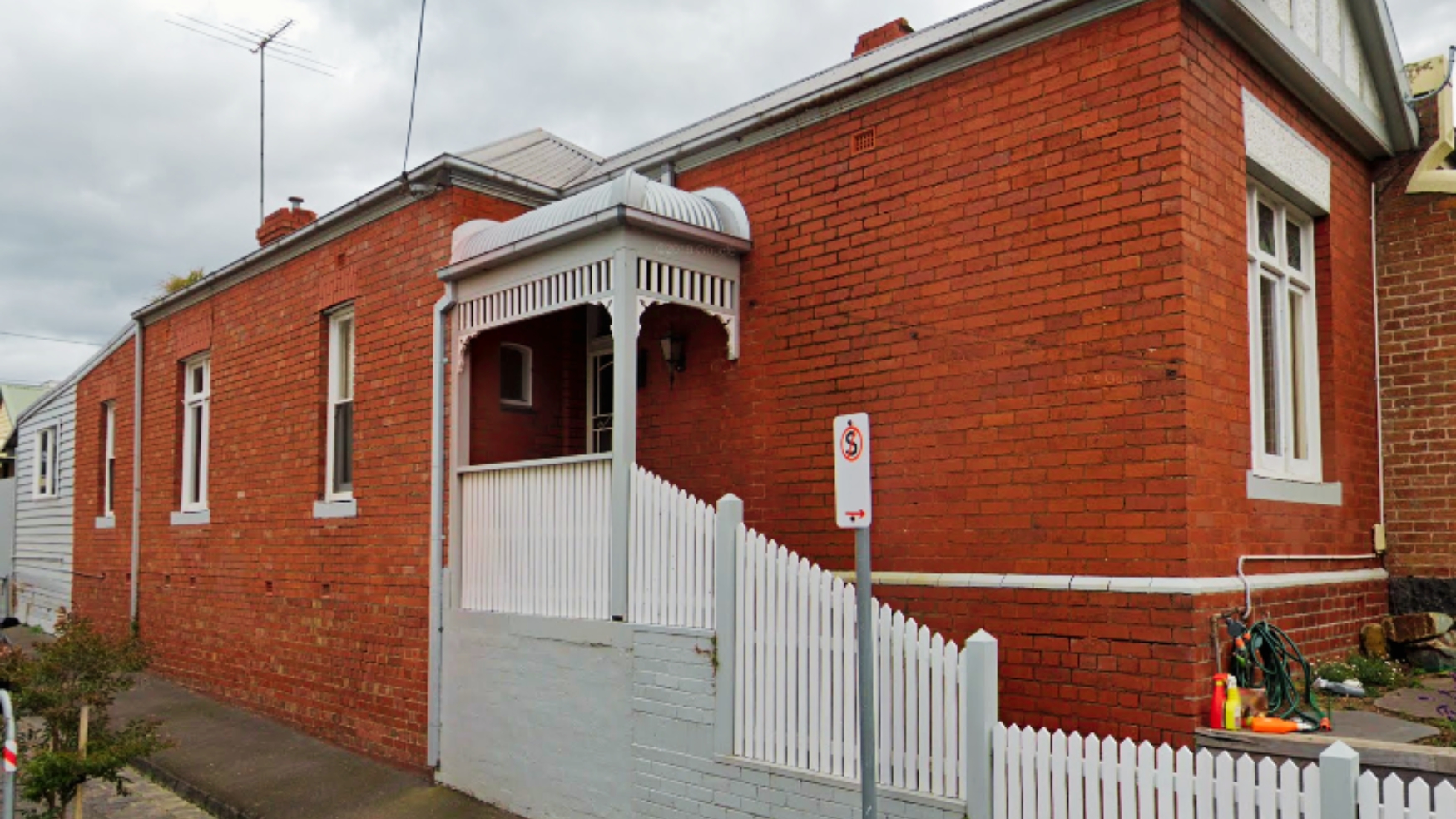 Red brick Clifton Hill home with painted timber verandah, decorative fretwork and a white picket fence